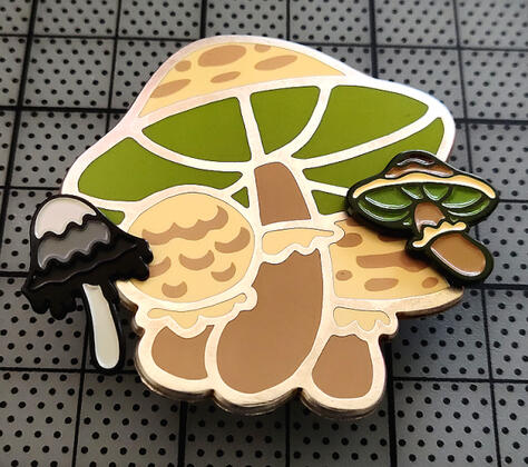 Mushroom Pins, a large hard enamel pin with silver metal and two smaller soft enamel pin with green and black metal.